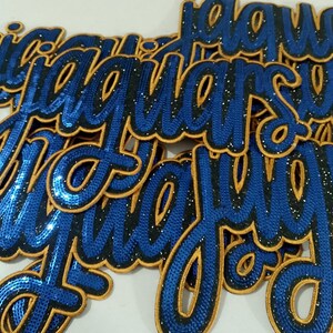 Puede incluir: Primer plano de varios apliques bordados en azul y dorado que deletrean la palabra "Jaguar" en una fuente cursiva. Los apliques tienen un aspecto texturizado con contornos dorados y una pizca de purpurina.