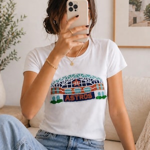 May include: White t-shirt featuring a colorful embroidered design of a stadium with the text "ASTROS". The shirt is paired with blue jeans. The person is holding a phone.