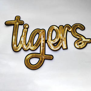 Puede incluir: Un parche dorado con lentejuelas con la palabra "tigers" en fuente cursiva. Las letras están delineadas en negro y rellenas de lentejuelas doradas. Ideal para ropa o accesorios.