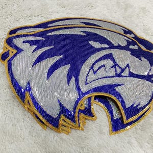 May include: A stack of blue and silver sequined patches in the shape of a snarling wildcat head. The patches have a gold border and are on a white, textured surface.