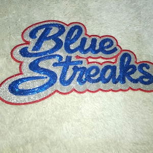 Pode incluir: Um patch brilhante com as palavras "Blue Streaks" em azul. O texto é delineado com glitter prateado, com uma borda vermelha. O patch está em uma superfície branca e texturizada.