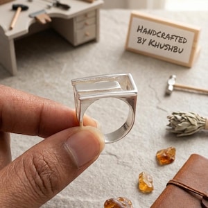 May include: A silver ring with a rectangular top, held in a hand. The ring is handcrafted, as indicated by the text "HANDCRAFTED BY KHUSHBU" on a small sign. The background includes jewelry-making tools and amber stones.
