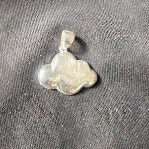 May include: A silver cloud-shaped pendant with a loop for a chain. The pendant has a smooth, polished surface and is set against a dark, textured background. The jewelry is a simple, elegant design.