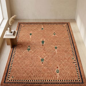 Puede incluir: Una alfombra rectangular con un patrón de ladrillos en tonos naranja y beige. La alfombra presenta figuras estilizadas en verde y blanco, con un borde negro y azul marino. La alfombra está colocada sobre un suelo de madera clara.