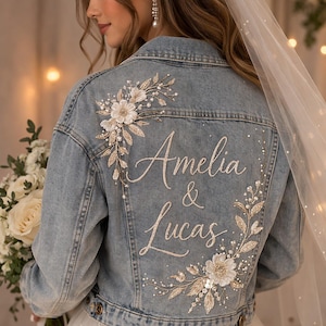 Könnte beinhalten: Hellblaue Jeansjacke mit floraler Stickerei und den Namen "Amelia & Lucas" in Schreibschrift. Die Jacke hat weiße und goldene Verzierungen, perfekt für eine Hochzeit oder einen Braut-Event.