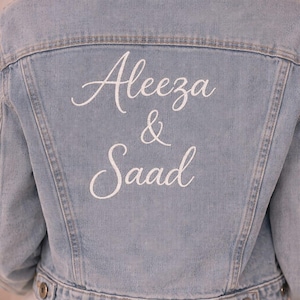 May include: Pale blue denim jacket featuring the embroidered names "Aleeza & Saad" in white script on the back. The jacket has a traditional design with button fastenings. Bridal wear.