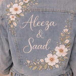 May include: Pale blue denim jacket with floral embroidery and the names "Aleeza & Saad" in white script. The jacket features white flowers, green leaves, and small pearl-like accents, ideal for a wedding or special event.