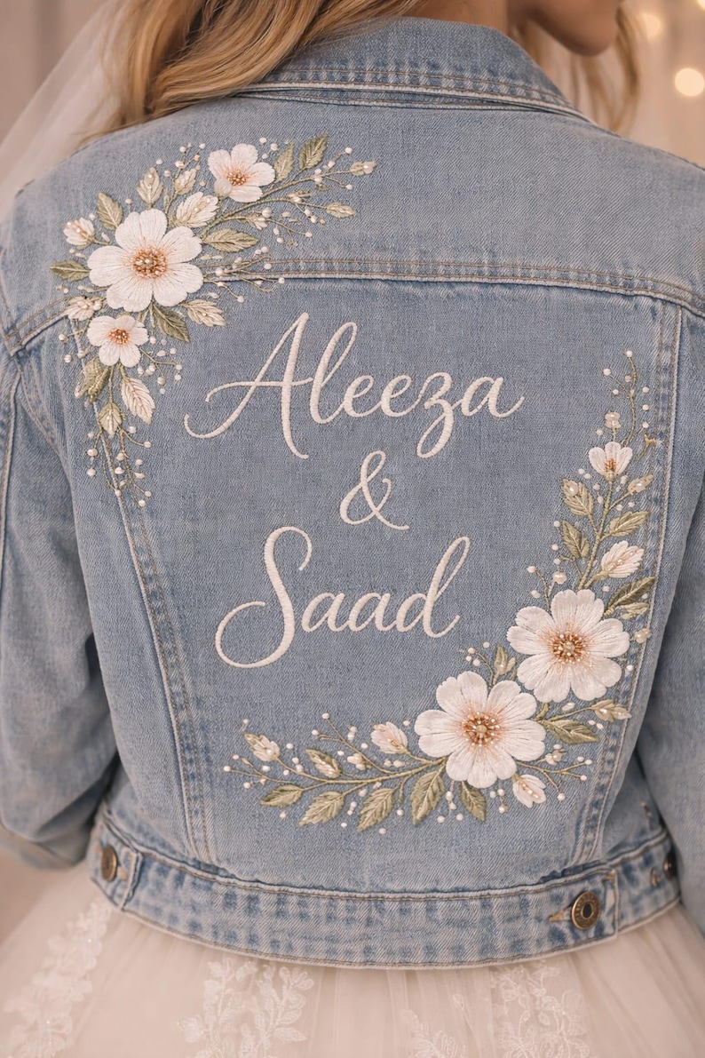 May include: Pale blue denim jacket with floral embroidery and the names "Aleeza & Saad" in white script. The jacket features white flowers, green leaves, and small pearl-like accents, ideal for a wedding or special event.
