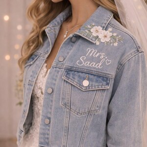 May include: Pale blue denim jacket with floral embroidery and the text "Mrs. Saad" in white script. The jacket has a classic button-up design and is worn over a white lace dress, ideal for a bride.