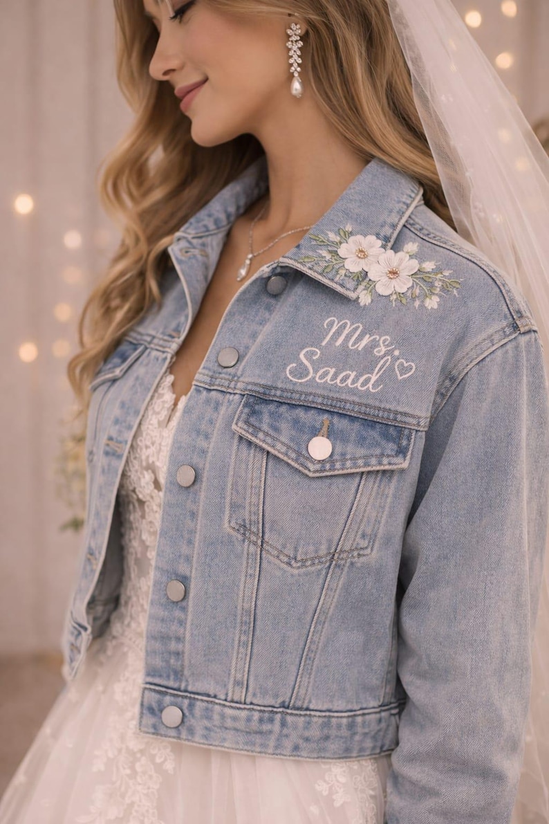 May include: Pale blue denim jacket with floral embroidery and the text "Mrs. Saad" in white script. The jacket has a classic button-up design and is worn over a white lace dress, ideal for a bride.
