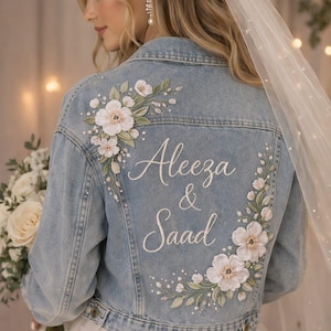 May include: Pale blue denim jacket with floral embroidery and the names "Aleeza & Saad" in white script. The jacket features white flower and green leaf designs, ideal for a wedding or bridal event.