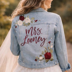 May include: Pale blue denim jacket with floral embroidery in white, burgundy, and green. The back of the jacket features the embroidered text "Mrs. Looney" in burgundy script. A bridal jacket for a wedding.