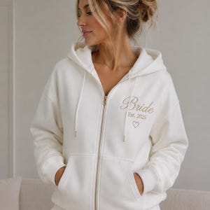 May include: A cream-colored zip-up hoodie featuring a hood with drawstrings, long sleeves, and two front pockets. The left chest area is embroidered with the word Bride in script, followed by the text Est. 2025 and a small heart outline.
