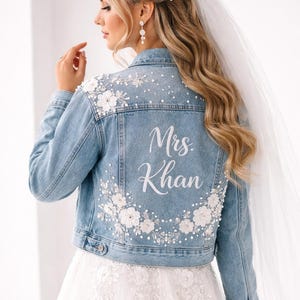 Peut inclure: Veste en jean bleu clair ornée de fleurs blanches et de perles. Le dos porte l'inscription "Mrs. Khan" en blanc. Une veste de mariée, parfaite pour un mariage.