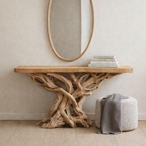 Puede incluir: Una consola de madera con base de raíz de árbol y tapa rectangular. Un espejo ovalado dorado cuelga sobre la mesa. Hay libros apilados sobre la mesa. Una manta gris descansa sobre un pequeño puf redondo tapizado.