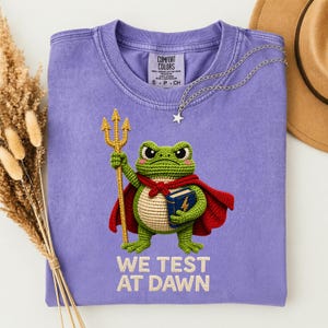 Op de afbeelding: Een paars Comfort Colors T-shirt met een gehaakte kikker-superheld die een drietand en een boek vasthoudt. De kikker draagt een rode cape en de tekst "WE TEST AT DAWN" staat eronder. Een zilveren ketting met een sterhanger ligt op het shirt.