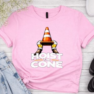 May include: A light pink t-shirt featuring the phrase "HOIST THE CONE" in white block letters. The graphic shows a traffic cone held by gloved hands. The cone is orange with white stripes.