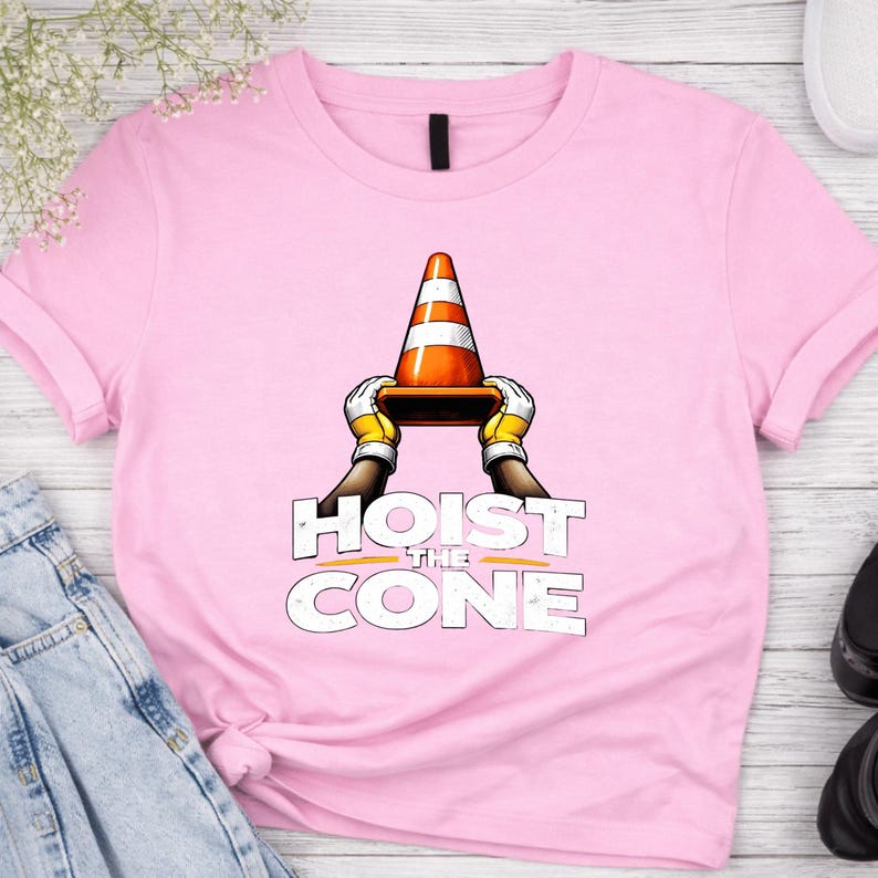 May include: A light pink t-shirt featuring the phrase "HOIST THE CONE" in white block letters. The graphic shows a traffic cone held by gloved hands. The cone is orange with white stripes.