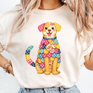 May include: A cream t-shirt with a cartoon dog design. The dog is decorated with a patchwork pattern in pink, yellow, blue, and green. It has a blue collar with a heart-shaped tag and a happy expression. The design is bright and cheerful.