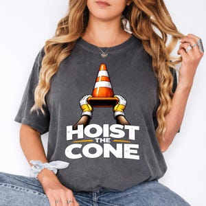 Pode incluir: T-shirt cinzenta escura com um gráfico de um cone de trânsito segurado por mãos com luvas, com o texto "HOIST THE CONE" em branco. O cone é laranja e branco. A t-shirt é de estilo casual.