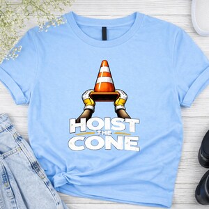 May include: Light blue t-shirt featuring a graphic of a traffic cone held by gloved hands, with the text "HOIST THE CONE" in a bold, distressed font. The design is centered on the shirt.