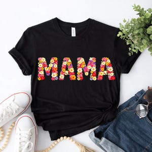 Könnte beinhalten: Schwarzes T-Shirt mit dem Wort "MAMA" aus bunten Blumen. Das Shirt hat einen Rundhalsausschnitt. Das Bild zeigt auch weiße Turnschuhe und blaue Jeans. Ein lässiges Oberteil für Mütter.