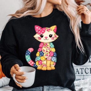 Pode incluir: Camisola preta com um design vibrante de gato patchwork. O gato apresenta uma cabeça rosa com bolinhas, detalhes florais e um corpo patchwork em vários padrões e cores. O gato é retratado sentado com a cauda enrolada.