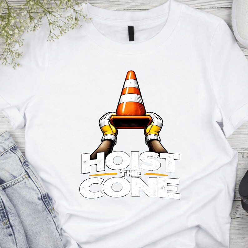 May include: White t-shirt featuring a graphic of a traffic cone held by gloved hands, with the text "HOIST THE CONE" in a bold, stylized font. The cone is orange and white, and the gloves are yellow.
