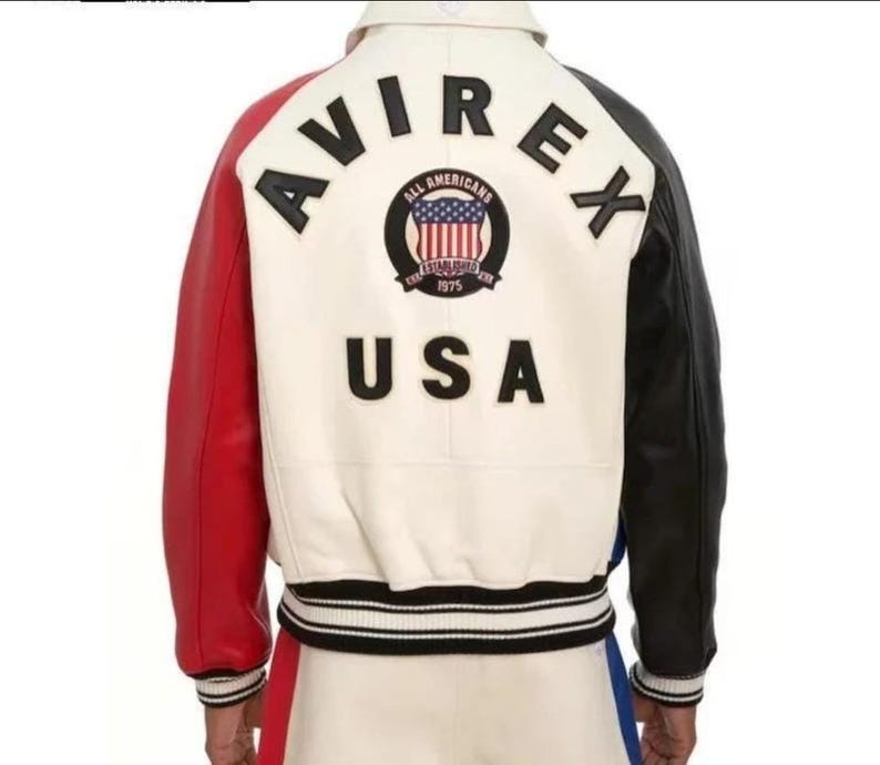 May include: A varsity jacket featuring a cream-colored back panel with the black text AVIREX and USA. A circular patch displays the text ALL AMERICANS ESTABLISHED 1975 with a US flag. The jacket has a red right sleeve and a black left sleeve.