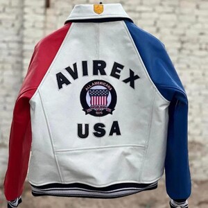 May include: A white leather varsity jacket with a red right sleeve and a blue left sleeve. The back features the black text AVIREX and USA, with a circular patch in the center displaying the text ALL AMERICANS ESTABLISHED 1975 and a US flag.