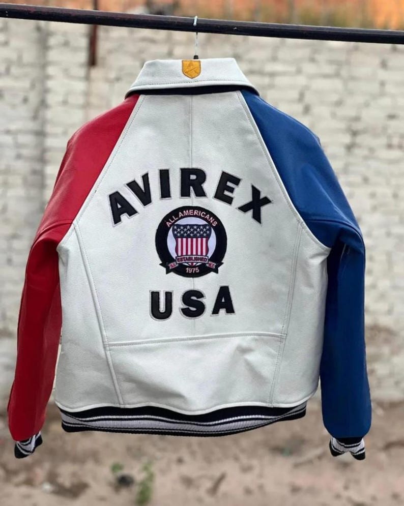 May include: A white leather varsity jacket with a red right sleeve and a blue left sleeve. The back features the black text AVIREX and USA, with a circular patch in the center displaying the text ALL AMERICANS ESTABLISHED 1975 and a US flag.