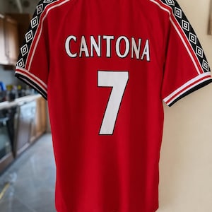 Pode incluir: Uma camisola desportiva vermelha de manga curta com gola branca e detalhes em preto e vermelho. As costas exibem o nome CANTONA e o número 7 em branco com contornos pretos. As mangas têm faixas pretas com padrões de losangos brancos.