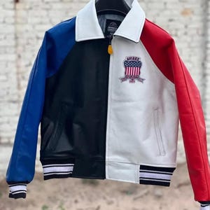 May include: A color-blocked jacket featuring a white collar, a blue left sleeve, a black front panel, and a white right front panel with a red sleeve. The chest displays an Avirex patch with a flag design. It includes a zipper closure and striped cuffs.