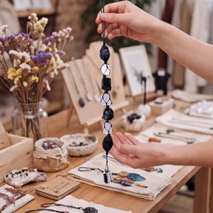 May include: A vertical hanging ornament featuring a series of alternating black and clear, translucent, irregularly shaped stones encased in a black knotted cord netting, ending in a small decorative tassel at the bottom of the structure.