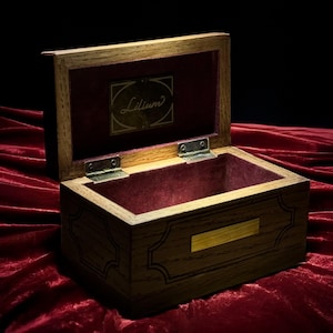 Può includere: Scatola portagioie in legno con coperchio aperto, che rivela un interno in velluto bordeaux. La scatola ha una forma rettangolare con intagli decorativi e una targhetta dorata. La parola "Lilium" è visibile all'interno del coperchio.