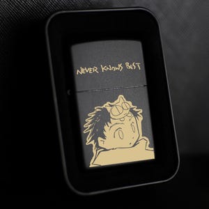 Puede incluir: Encendedor de metal negro con el texto "NEVER KNOWS BEST" y una ilustración de un personaje de dibujos animados. El encendedor está en un estuche rectangular negro.