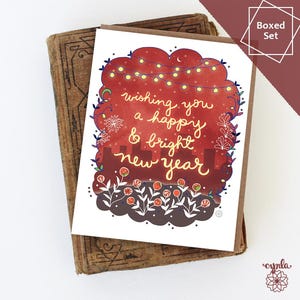 May include: A boxed set of greeting cards with a festive design. The card features the text "wishing you a happy & bright new year" in a handwritten style, surrounded by decorative elements like lights and flowers. The card is set on a vintage book.