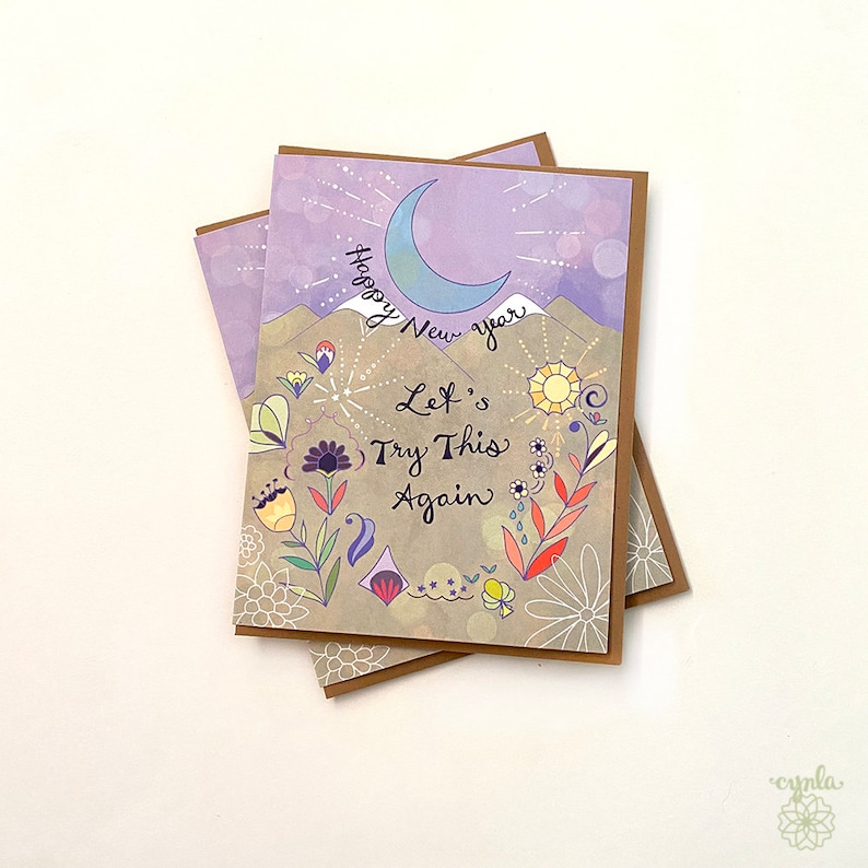 May include: A greeting card featuring a light purple sky with a blue crescent moon, tan mountains, and colorful floral illustrations. The text reads Happy New Year and Let's Try This Again in black script. The card is paired with a brown envelope.