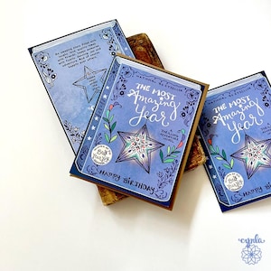 May include: Greeting cards with a book theme. The cards feature a blue cover design with the text "The Most Amazing Year" and "Happy Birthday". The cards have a star and floral design.