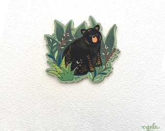 May include: A black bear with gold and white markings sits in a green leafy forest. The bear has a brown nose and is looking directly at the viewer.