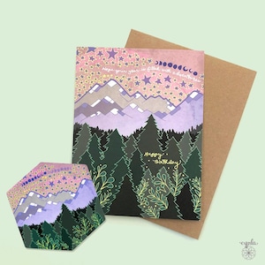 May include: A birthday card and coaster set featuring a mountain and forest scene under a starry sky. The card reads "Hope your year is filled with adventures" and "Happy Birthday". The coaster has a similar design.