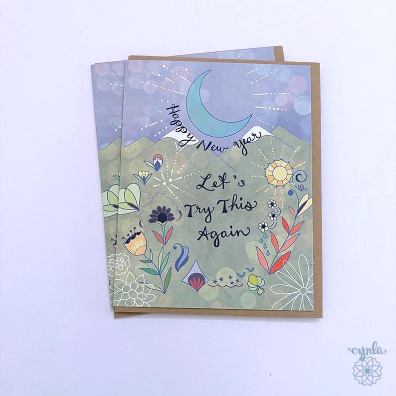 May include: A greeting card featuring a light blue crescent moon, mountains, and colorful floral illustrations. The text reads Happy New Year and Let's Try This Again in black script. The card is set against a light purple and green background.