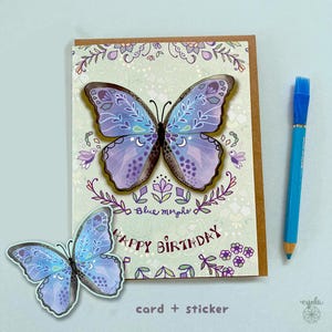 May include: A greeting card and a matching sticker featuring a blue and purple butterfly with white crescent moon and floral patterns. The card displays the text "Blue morpho" and "HAPPY BIRTHDAY" in dark purple. A blue pencil is placed to the right.