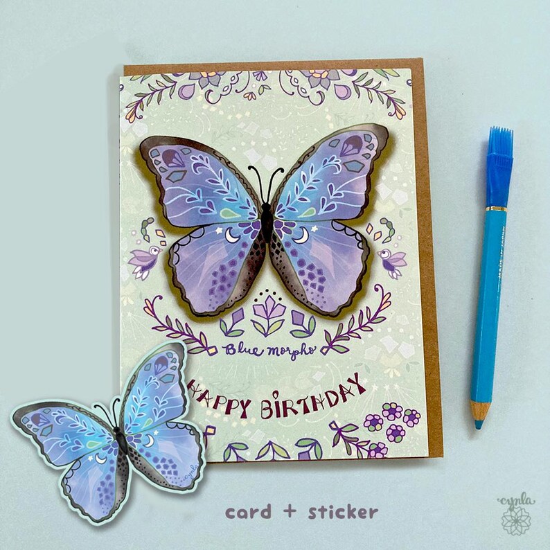May include: A greeting card and a matching sticker featuring a blue and purple butterfly with white crescent moon and floral patterns. The card displays the text "Blue morpho" and "HAPPY BIRTHDAY" in dark purple. A blue pencil is placed to the right.