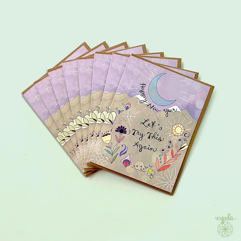 May include: A stack of greeting cards featuring a lavender sky, a blue crescent moon, and tan mountains. The text reads Happy New Year Let's Try This Again. The design includes colorful floral illustrations, a sun, and starburst patterns.