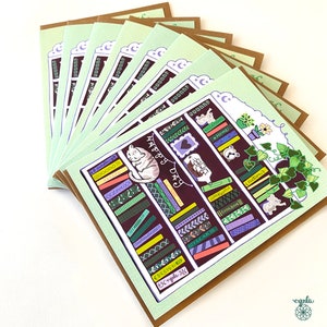 May include: A stack of greeting cards with a whimsical bookshelf design. Each card features a detailed illustration of a bookshelf with books, a sleeping cat, and the words "Happy Day". The cards have a light green border and a brown backing.