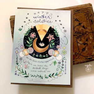 Puede incluir: Una tarjeta ilustrada azul y dorada con una luna creciente, el sol y flores. El texto dice "Feliz Solsticio de Invierno" y "Cuando las primeras estrellas aparecieron, supimos que los d&iacute;as m&aacute;s oscuros hab&iacute;an quedado atr&aacute;s".