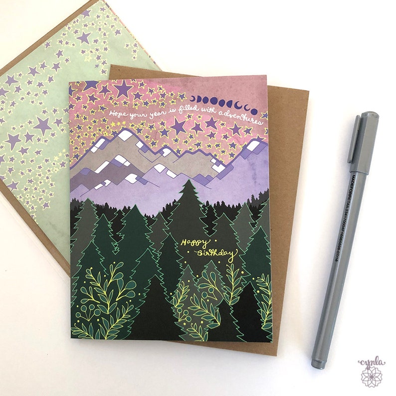 May include: A birthday card with a forest scene. The card features a purple and pink sky with stars and a crescent moon. The text on the card reads "Hope your year is filled with adventures. Happy Birthday."