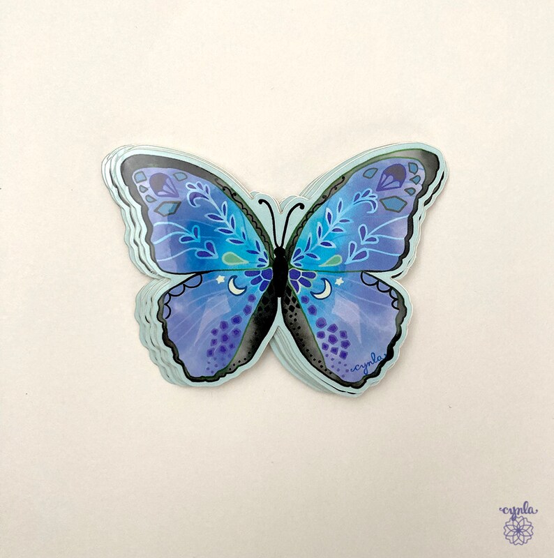 May include: A stack of die-cut butterfly stickers featuring a blue and purple gradient color scheme. The wings are decorated with white leaf patterns, small crescent moons, and stars. The body is black, and the word cynla is printed in small text.