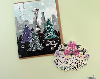 May include: A rectangular greeting card featuring a Seattle skyline illustration with the Space Needle, stylized evergreen trees, snowflakes, and the text Happy Holidays, alongside a die-cut sticker with pink flowers and the word Washington.